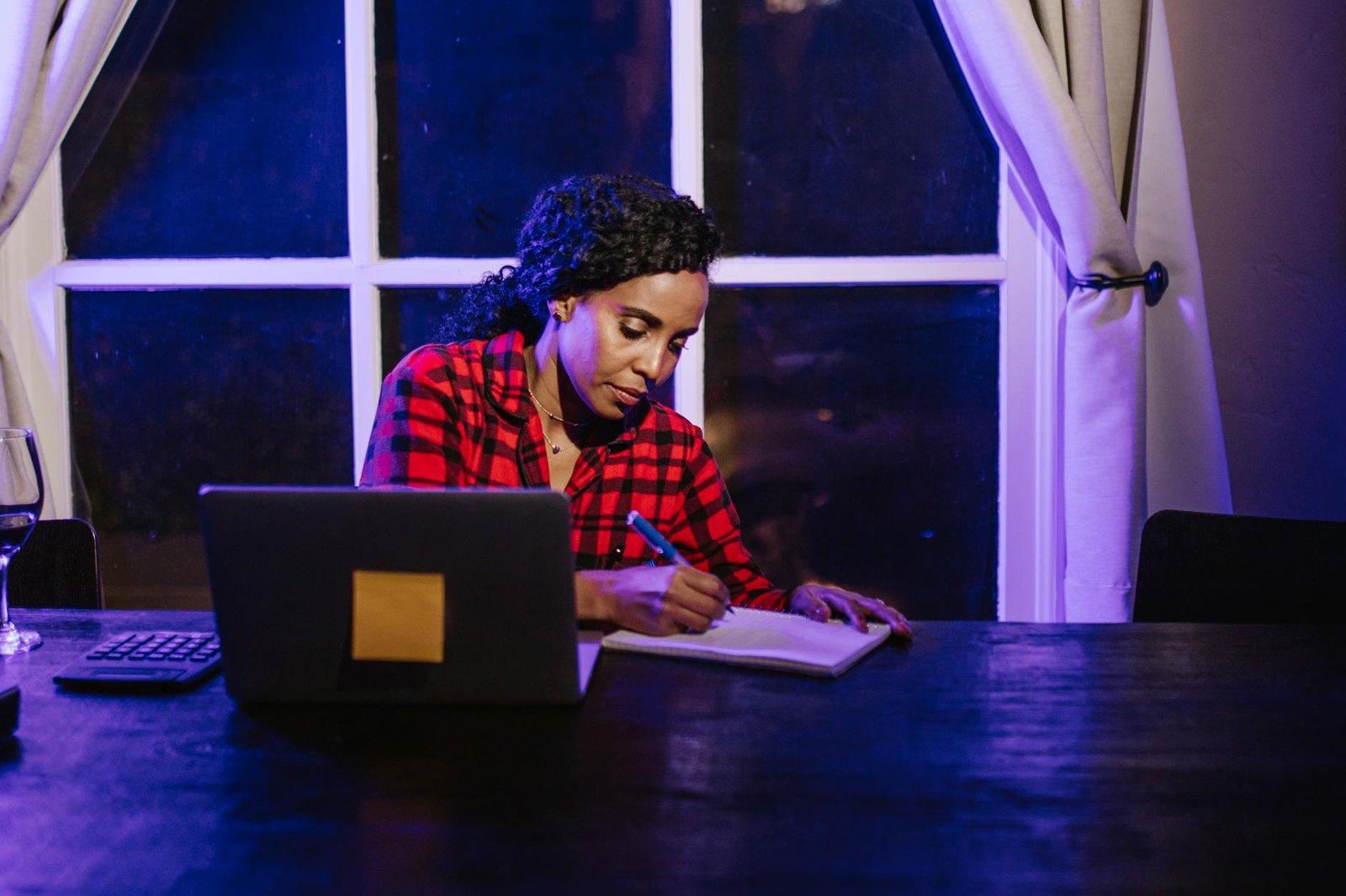 A woman in a plaid shirt writing in a notebook at a home workspace at night.