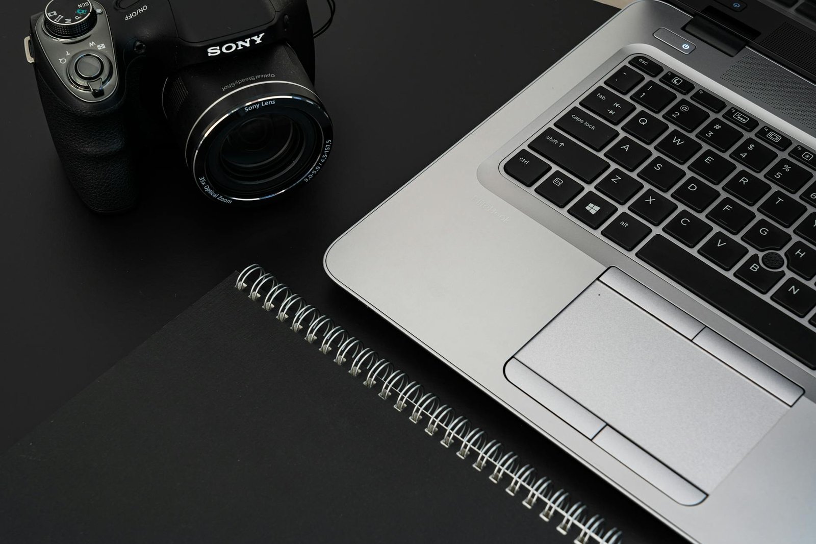 A stylish office desk featuring a laptop, camera, and spiral notebook on a dark surface.