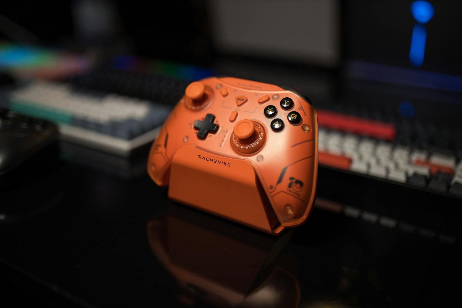 Orange gaming controller on black desk with colorful backlit keyboard in background.
