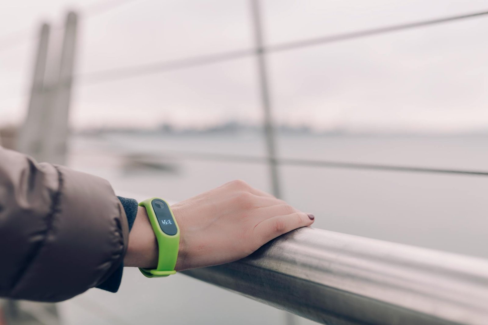 A woman wearing a green smartwatch rests her hand on a railing with a blurred city skyline in the background.