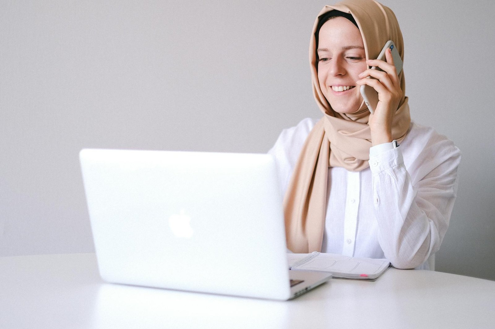 Smiling woman in a hijab uses laptop and phone at a home office.