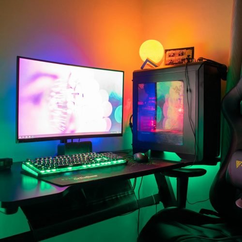 A colorful gaming setup featuring a PC, monitor, and gaming chair with RGB lights in a dark room.