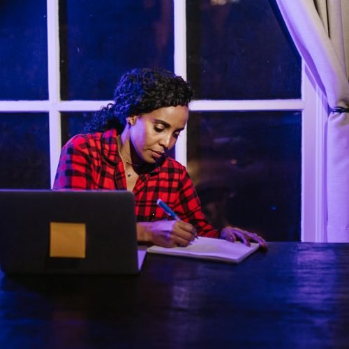 A woman in a plaid shirt writing in a notebook at a home workspace at night.