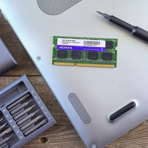 Close-up of an open laptop with RAM module, screwdriver, and toolset on a wooden surface.