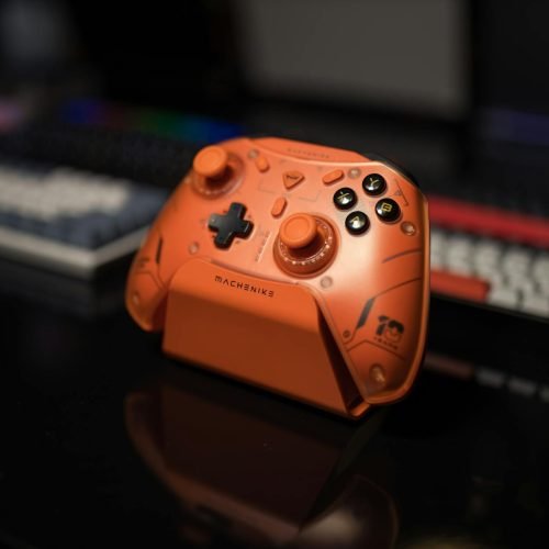 Orange gaming controller on black desk with colorful backlit keyboard in background.