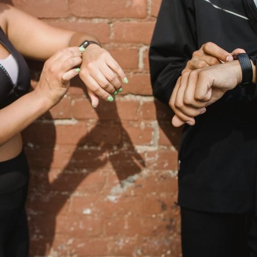 Two people in sportswear checking their fitness trackers outdoors against a brick wall.