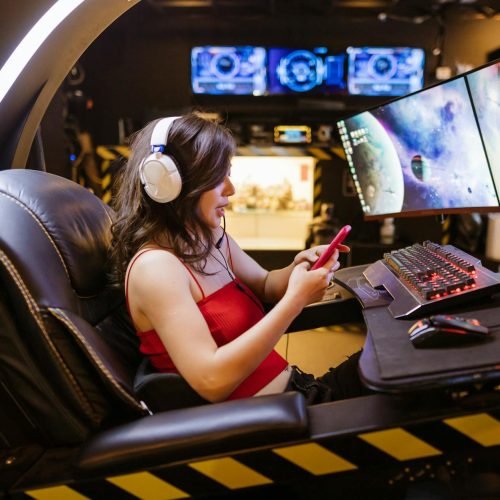 Young woman enjoying computer gaming at home with a high-tech gaming setup and smartphone.