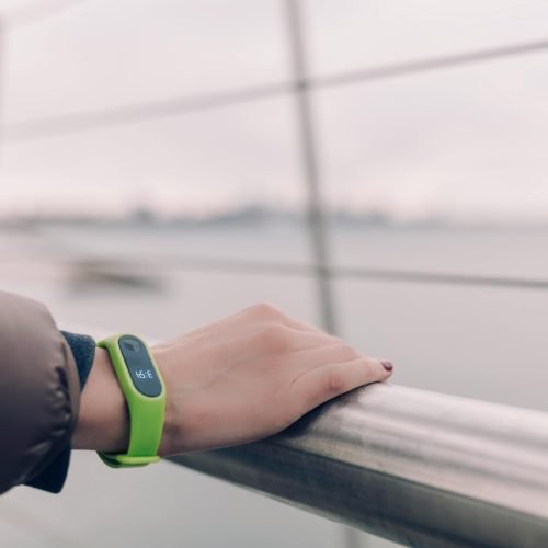 A woman wearing a green smartwatch rests her hand on a railing with a blurred city skyline in the background.