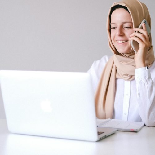 Smiling woman in a hijab uses laptop and phone at a home office.