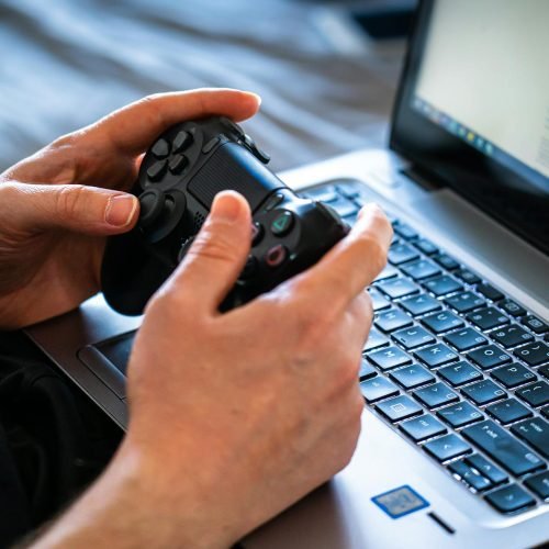 Person holds a gaming controller while interacting with a laptop, emphasizing modern gaming technology.