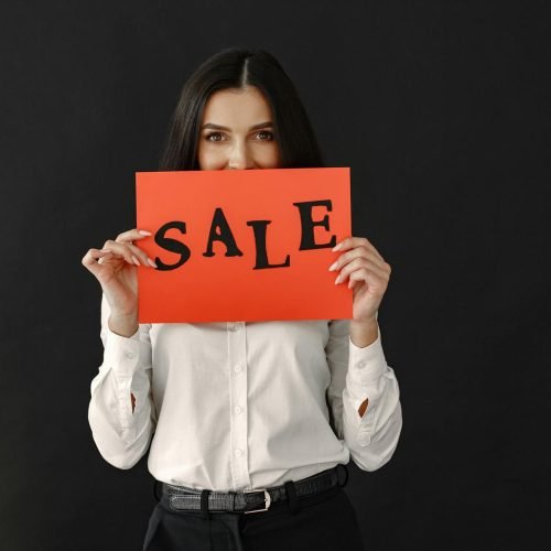 Woman in white shirt holding a red sale sign against a black backdrop. Ideal for advertising.